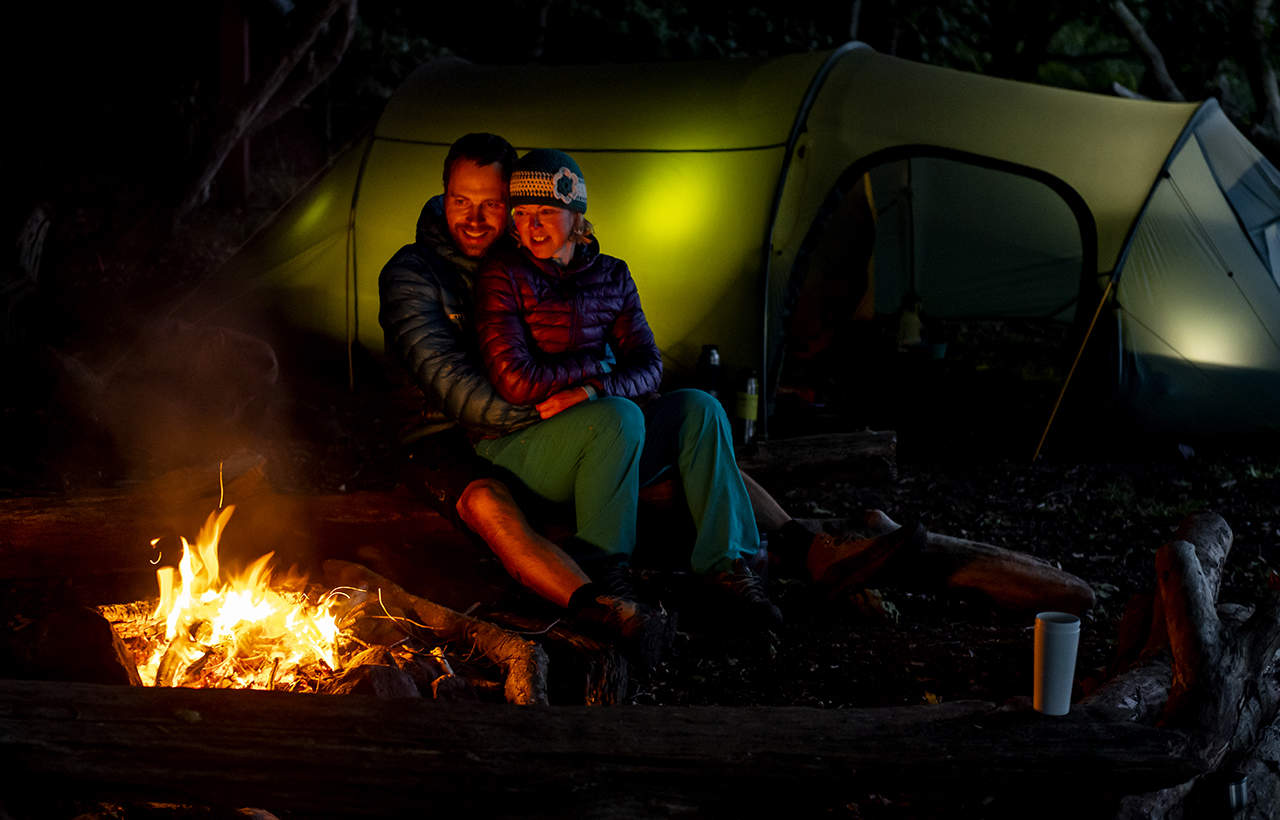 Erik B. Jørgensen og Tine Henriksen sidder op ad hinanden ved bålet, telt i baggrund. Titel: Hygge i den danske natur, med Erik B. Jørgensen. Foto Rene Schütze