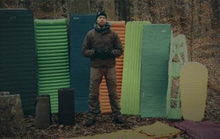 Liggeunderlag, jeg bruger, til friluftsliv. Erik B. Jørgensen med mange liggeunderlag stående bag sig, i en skov.