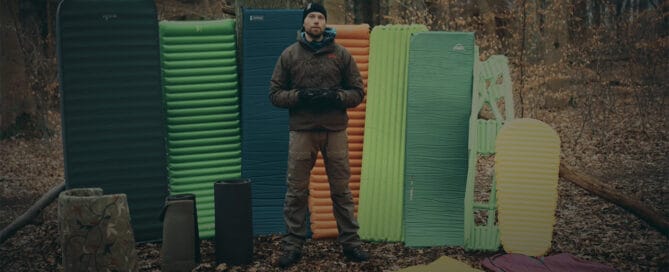 Liggeunderlag, jeg bruger, til friluftsliv. Erik B. Jørgensen med mange liggeunderlag stående bag sig, i en skov.