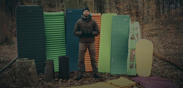 Liggeunderlag, jeg bruger, til friluftsliv. Erik B. Jørgensen med mange liggeunderlag stående bag sig, i en skov.